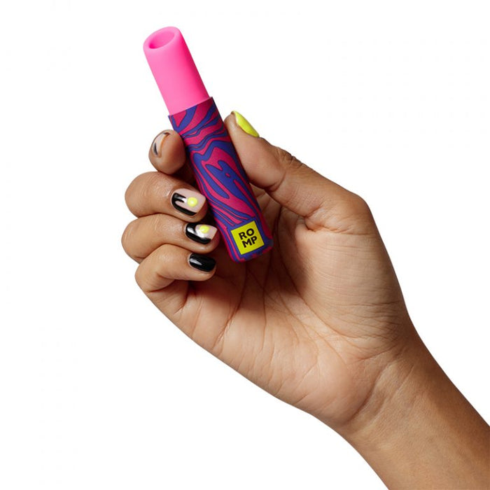 A hand with manicured nails painted in black, white, and yellow holds a small, colorful clitoral stimulator. The object has a pink tip and a blue and red wavy patterned body with a yellow label that reads "ROMP." This is the Romp Lipstick First Time Pleasure Air Clitoris Stimulator Vibrator by WOW, specifically designed to deliver heightened clitoral orgasms through Pleasure Air Technology.