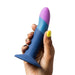 A hand with colorful painted nails is holding a blue, pink, and purple silicone dildo with a flared base. This sleek toy, perfect for anal play, features a smooth design and the product name "Romp Piccolo First Time 5 Inch Silicone Dildo" from the brand WOW embossed on it.