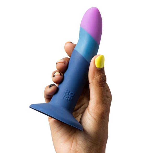 A hand with painted nails holds a blue, teal, and purple silicone dildo with a suction base from the Lovehoney Romp Piccolo First Time Pegging Kit - Strap on Harness + Dildo. The product has the word "ROMP" embossed on the side.