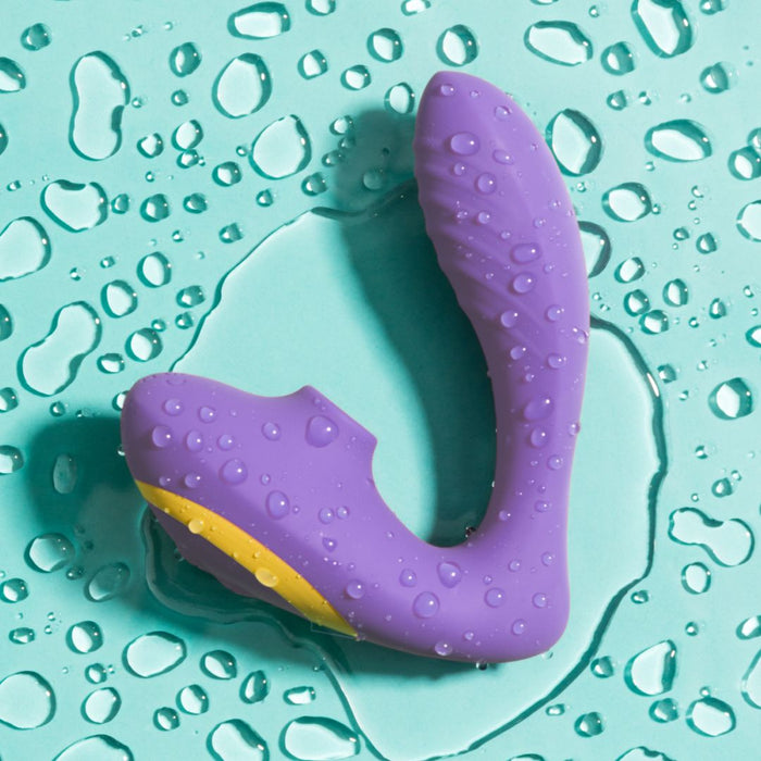 A purple silicone Romp Reverb Dual Stimulation Pleasure Air Vibrator with a yellow accent from the brand WOW rests on a teal waterproof surface. Water droplets are scattered around and on the device, indicating it is water-resistant or waterproof and ready for clitoral suction stimulation.