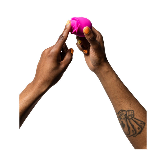 A person holds a small, pink, geometric-shaped object between their hands. One hand has fingers pointing at it. The person has a tattoo of four ribbons on their forearm. The background is white, subtly highlighting the innovative Romp Rose Pleasure Air Clitoris Stimulator - Pink by WOW that they are showcasing.