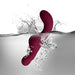 The Ruby Glow Blush External Grinding and G-Spot Vibrator with Remote by Rocks Off, in a deep burgundy hue, is shown partially submerged in water. Splashes and droplets surround the curved, waterproof device, emphasizing its water-resistant capabilities against a white and minimalistic background.