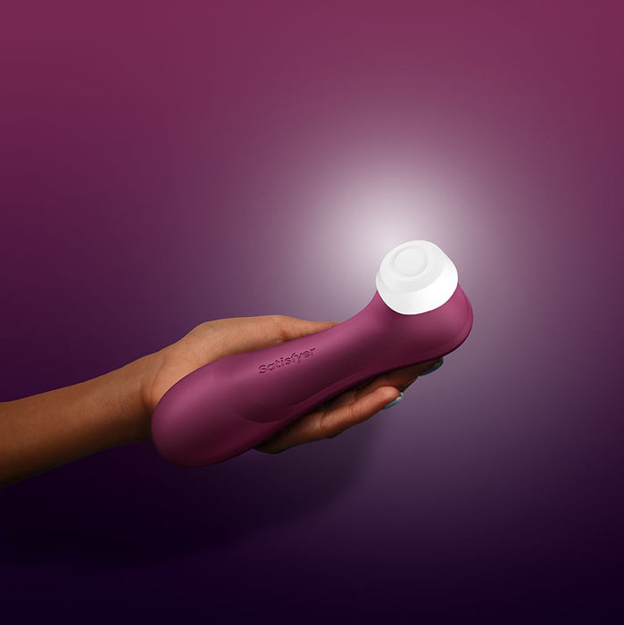 A hand holds a wine red Satisfyer Pro 2 Gen 3 App Controlled Pressure Wave Clitoral Stimulator, a personal pleasure device known for its clitoral suction and air pulsation technology, against a gradient background ranging from deep purple to light purple. The device features a white silicone head at its end.