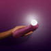 A hand holds a wine red Satisfyer Pro 2 Gen 3 App Controlled Pressure Wave Clitoral Stimulator, a personal pleasure device known for its clitoral suction and air pulsation technology, against a gradient background ranging from deep purple to light purple. The device features a white silicone head at its end.