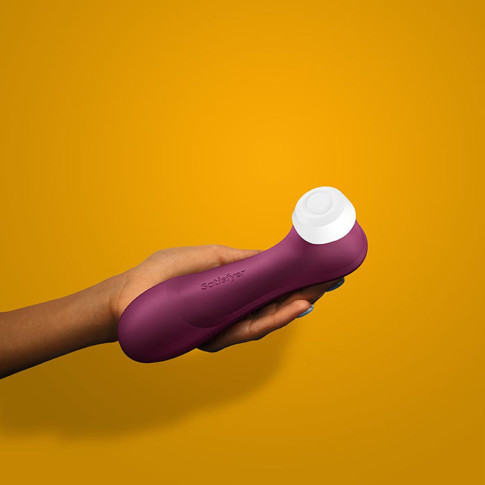 A person holds the Satisfyer Pro 2 Generation 3 Pressure Wave Clitoral Stimulator by Satisfyer, featuring a soft matte finish and a white circular attachment, against a solid orange background. The wine red, waterproof device is ergonomically curved for enhanced comfort.