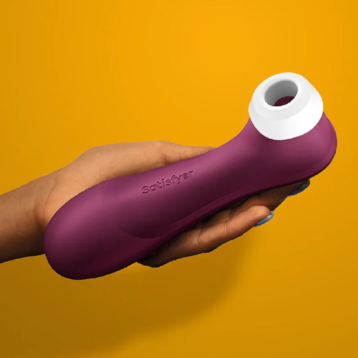 A hand is holding a wine-red Satisfyer Pro 2 Generation 3 Pressure Wave Clitoral Stimulator by Satisfyer, featuring white tips and designed for clitoral stimulation using air pulsation technology, against a solid mustard yellow background. The product boasts a sleek, ergonomic shape with the brand name engraved on its side.