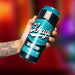 A tattooed hand holds a can of Schag's Sultry Stout Self-Lubricating Penis Stroker by Blush, known for its ultra discreet teal and black design with white cursive branding. The can boasts "Superb Crafted. Leisure Brew. 7.2% Alc/Vol. Only The Finest." The background is a blur of vibrant, warm colors.