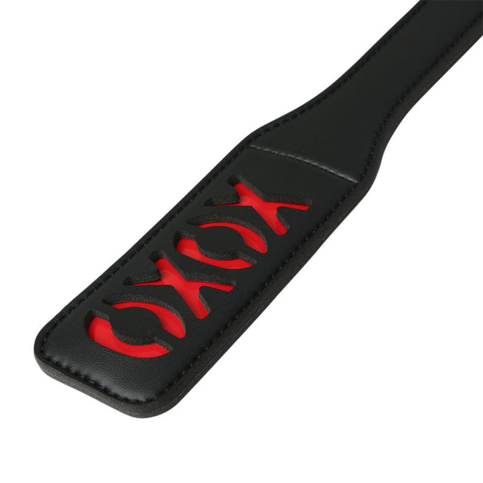 The Sportsheets Sex and Mischief XOXO Paddle Black 12 Inches features the word "XOXO" cut out, revealing a striking red backing underneath. This black vinyl paddle has a rectangular shape with rounded edges, making it an eye-catching BDSM accessory.