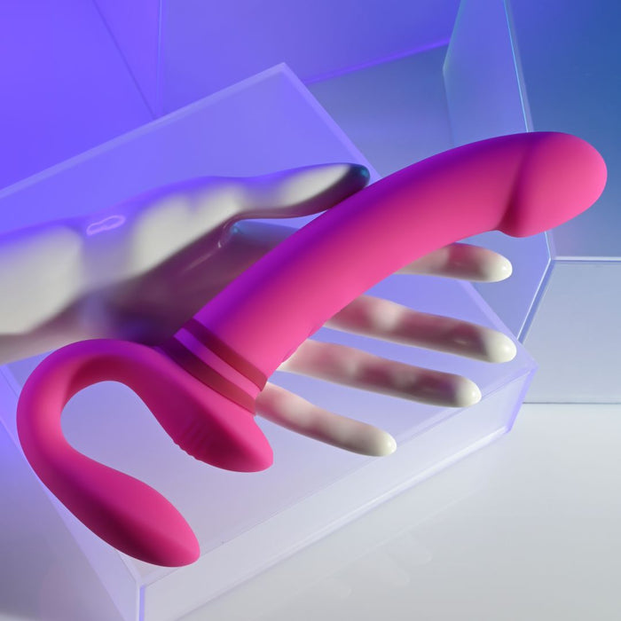 A hand holding the "Sharing Is Caring Double Ended Wearable Strapless Strap On Vibrator - Pink" from Evolved Novelties, made from phthalate-free silicone with a curved handle. The background features geometric shapes and a gradient of blue and purple hues. The toy's surface appears smooth and slightly reflective.