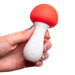 A hand is holding the Shroomie Mushroom Shaped Mini Wand Vibrator by Maia Toys, which features a red cap and a white stem. The vibrator appears to have a smooth texture and small buttons on the stem, suggesting its vibrating capabilities. The person's nails are painted light blue.