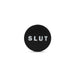 A black circular pin with the word "SLUT" written in bold white letters in the center, perfect for those who enjoy flaunting their playful side like the daring Blush Slut Silicone Butt Plug - Black.