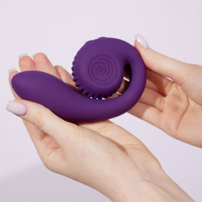 A person holds the Snail Gizi Compact Ultra Powerful 2 Motor Dual Stimulating Vibrator by Snail Vibe with both hands, displaying its unique and ergonomic design. The purple, curved, spiral-textured vibrator appears to be made of a soft material, likely silicone. The plain white background highlights the vibrator's distinctive features perfectly.