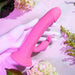 A white mannequin hand holds the Evolved Novelties' "Somebunny to Love Uncut Look Rabbit Vibrator," a pink dual-ended toy, on a light pink surface adorned with green leaves and white flowers. The vibrator features a larger main shaft alongside a smaller, rabbit-shaped attachment, all made from premium silicone.