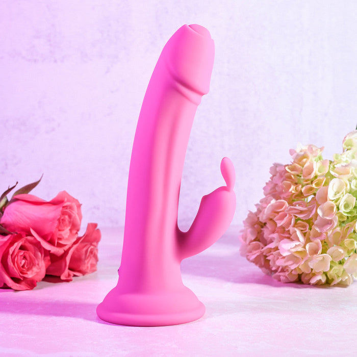 The Somebunny to Love Uncut Look Rabbit Vibrator by Evolved Novelties, featuring a pink silicone exterior with powerful motors, a curved shaft, and a smaller curved extension, is displayed upright against a light pink background. On the left side of the toy is a bouquet of pink roses, while on the right side is a cluster of green and pink hydrangeas.