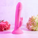 An Evolved Novelties' "Somebunny to Love Uncut Look" Rabbit Vibrator, in a soft pink color, stands upright on a matching surface. The powerful motor and phallic-shaped silicone design include a smaller attachment at the base. In the background, softly lit pink roses and light-colored hydrangeas frame the scene.