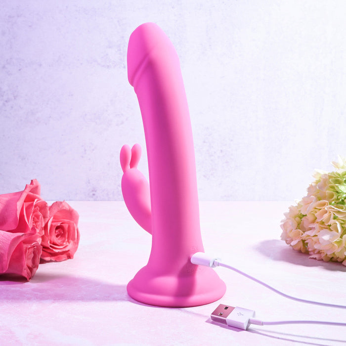 A pink silicone Somebunny to Love Uncut Look Rabbit Vibrator by Evolved Novelties stands upright on a light pink surface, propped against a backdrop of roses on the left and a bunch of light green hydrangeas on the right. A USB charging cable is attached to its base. This waterproof rabbit vibrator features powerful motors for enhanced pleasure.