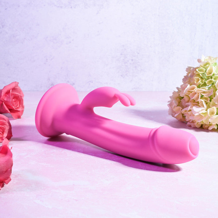 The Somebunny to Love Uncut Look Rabbit Vibrator by Evolved Novelties, crafted from premium silicone, features an insertable shaft, a curved clitoral stimulator, and a suction base. It lies on a light pink surface, accented by a bouquet of white and pink flowers. The powerful motors promise an unparalleled experience.