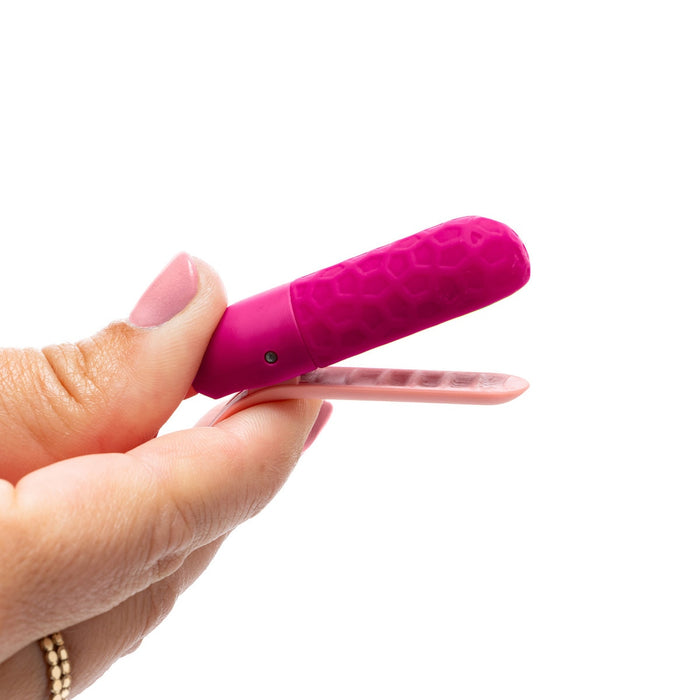 A hand holds a pink hair clip with a textured surface, its smooth prongs partially open like precision tools. The person's light pink nails resemble the gentle allure of the Sphinx Bluetooth App-controlled Vibrating Nipple Clamps.