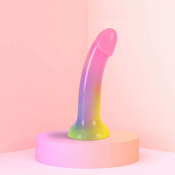 Displayed on a round, pink platform with a soft pink background is the Stargazer Rainbow Ombre Glitter 7 inch Silicone Dildo by Lovely Planet. Featuring a vibrant gradient of pink, blue, and yellow hues, this colorful dildo has a smooth, curved shape perfect for g-spot play and includes a flared base for added safety.