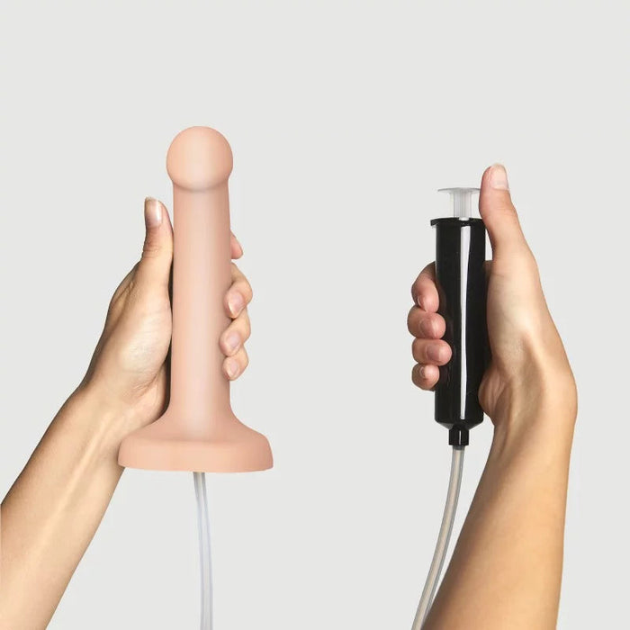 A pair of hands demonstrating the **Strap-On-Me 7.5 Inch Silicone Squirting Cum Dildo - Vanilla** by **Lovely Planet** held in the left hand, while the right hand holds a black pump device with an attached tube. Ideal for strap-on play, the setup is shown against a plain white background.