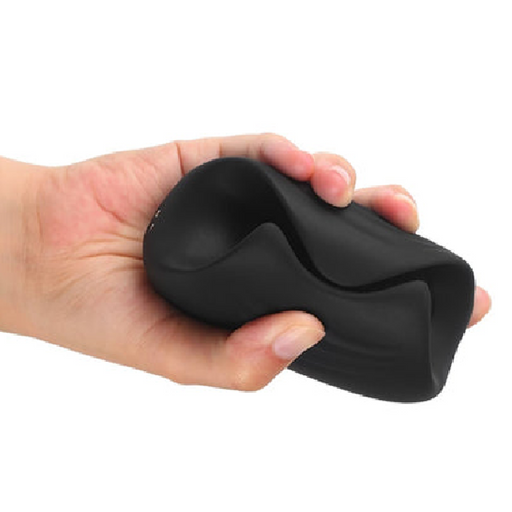 A hand squeezes the Sub One Tapping Penis Stroker and Frenulum Teaser, a black, flexible silicone object, against a white background. This durable item is 100% waterproof and highly versatile.