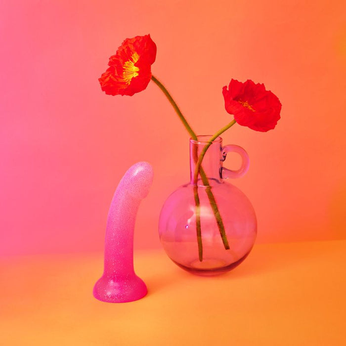 A Sunrise Pink Ombre Glitter 7-inch Silicone Dildo by Lovely Planet stands on a yellow surface next to a clear glass vase containing two vibrant red poppies. The background is a gradient of warm colors, transitioning from pink at the top to orange at the bottom.