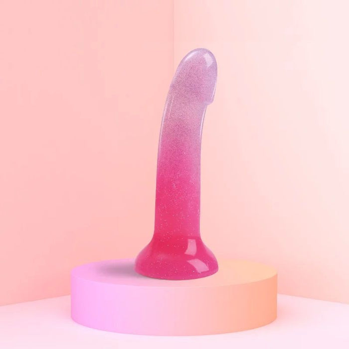 The Lovely Planet Sunrise Pink Ombre Glitter 7 inch Silicone Dildo, featuring a pink and purple gradient, is displayed standing upright on a pink circular platform against a softly lit pink background. The toy boasts a smooth, shiny surface with a slight curve and a flared base, making it ideal for prostate or g-spot play.