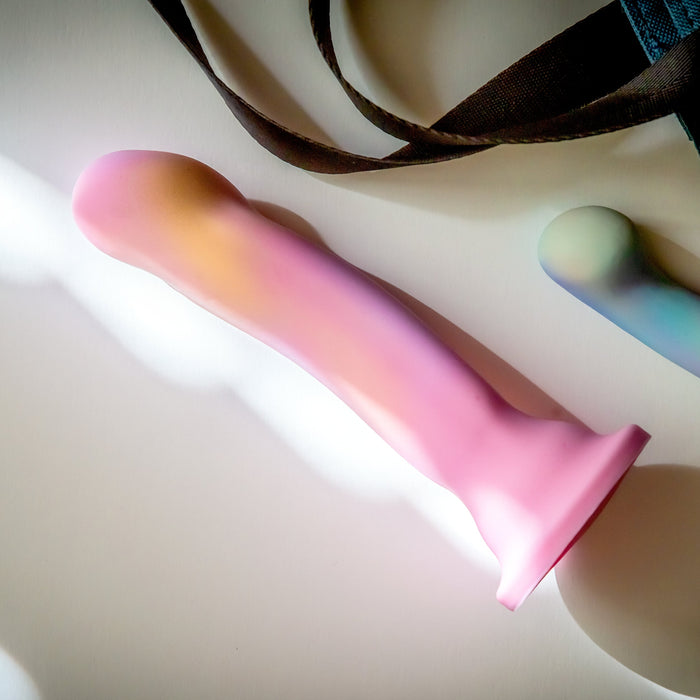 A silky smooth Sun's Out Pink Ombre 7.5 Inch G Spot Dildo by Blush, featuring a gradient of pink, purple, and yellow hues, lies on a white surface. Beside it is a fabric strap and there's a partial view of another similar object in the frame.