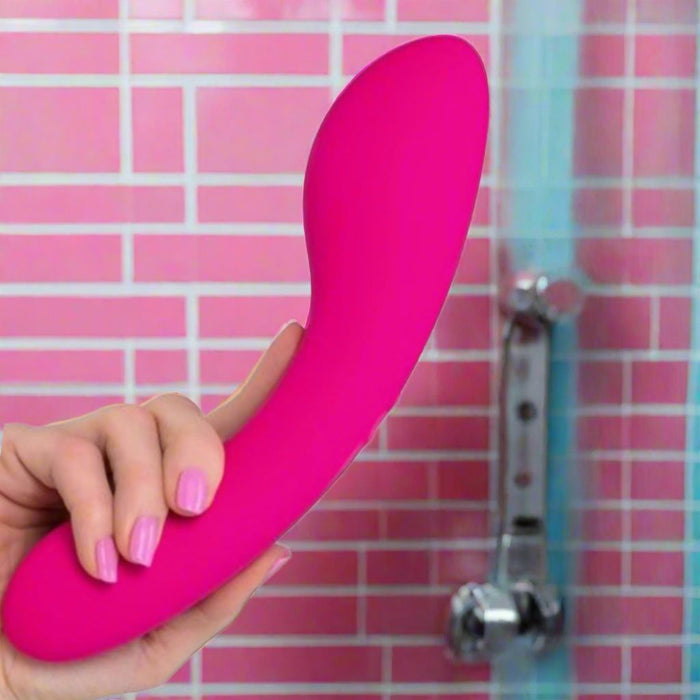 A hand with pink-painted nails holds the Swan Wand Powerful Rumbly Double Ended 2 Motor Vibrator by BMS Enterprises, made of 100% silicone, against a pink-tiled bathroom wall. A metal shower fixture is visible in the background.