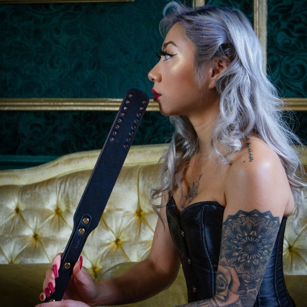 A person with long, wavy silver hair and an intricate tattoo on their arm holds a black paddle from the Temptasia Safe Word Bondage Kit with Suitcase by Blush, kissing it. Dressed in a black corset, they sit on a luxurious yellow velvet couch with green wallpaper in the background. The durable vegan leather glistens under the ambient light.