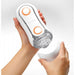 A person is holding a Tenga Flip Orb Discreet Penis Masturbator - Orange Crash, a cylindrical white and orange device from Tenga, in one hand and removing its white cap with the other hand. The device features a transparent section revealing pleasure orbs inside. The background is plain white.