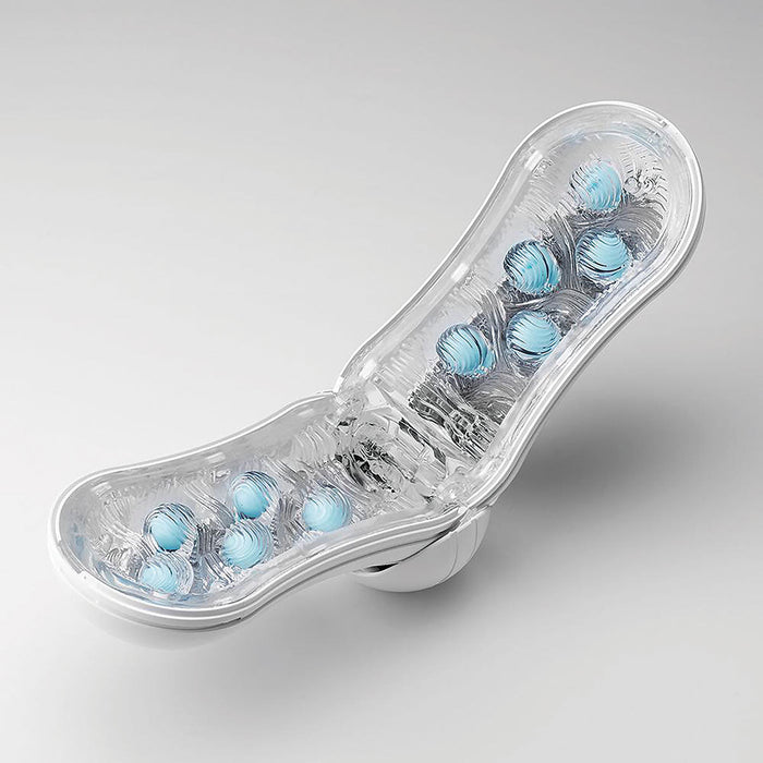 A clear, curved plastic case containing multiple blue, spherical pleasure orbs positioned in rows inside the open sections holds the Tenga Flip Orb Penis Masturbator - Blue Rush. The transparent lid reveals the contents clearly. The product is placed on a neutral, light-colored surface.