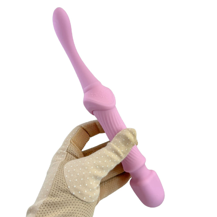 A hand wearing a beige, polka-dotted glove holds The Goddess Double Ended Wand + Pinpoint Vibe with 3 Attachments in pink by Nasstoys. The massager features a curved, narrow end for targeted stimulation and a larger, rounded end on the opposite side.