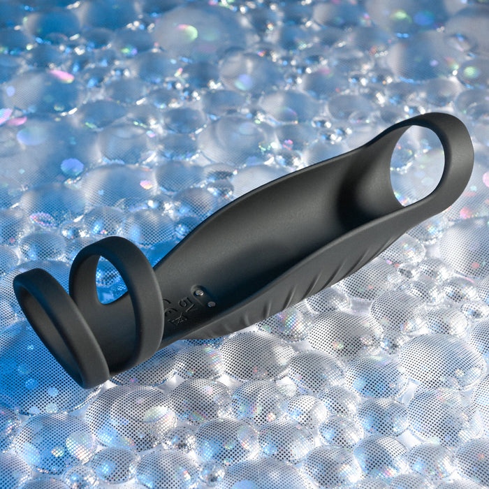 A stylish black item called The Rocketeer Vibrating Penis Enhancer Sheath by Evolved Novelties, consisting of body-safe silicone with a ring at each end, rests on a glittery, textured surface. This flexible cock sheath showcases subtle ridges along one side and boasts a matte finish, forming an intriguing design that doesn’t reveal its purpose at first glance.