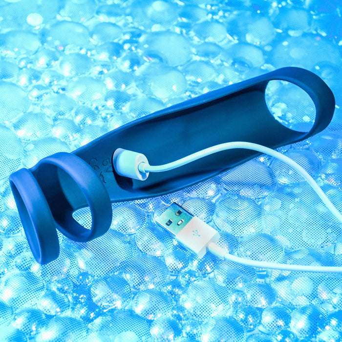 A black, ring-shaped vibrating penis enhancer sheath called "The Rocketeer" by Evolved Novelties, made of body-safe silicone and featuring a USB charging cable, set against a background of blue bubbles.