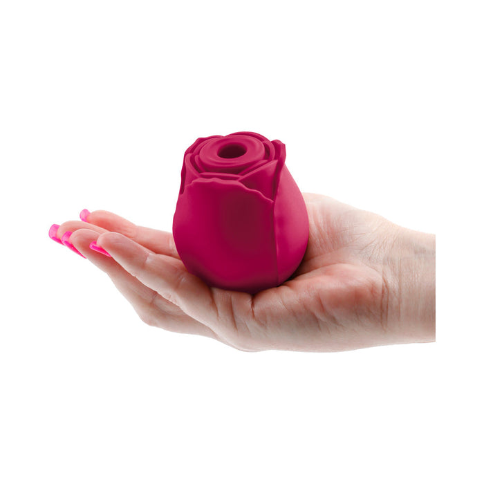 A hand with pink-painted nails holds The Rose Powerful Clitoral Air Pulsation Vibrator by NS Novelties, a small air pulsation device shaped like a rose in a deep red or burgundy color, against a white background. The Rose has a circular opening at the center designed for clitoral stimulation.