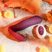 The Velvet Hammer Thrusting Hands-Free Dual Stimulation Vibrator from Evolved Novelties, a purple curved waterproof silicone device with a wireless remote control featuring plus, minus, and power buttons, lies on a textured surface surrounded by orange maple leaves and yellow flowers. The scene is decorated with an autumnal theme.