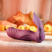 The Velvet Hammer Thrusting Hands-Free Dual Stimulation Vibrator by Evolved Novelties, an ergonomically designed silicone toy in a rich purple hue, rests on a textured surface set against a warm, autumn-themed backdrop adorned with scattered leaves and yellow flowers. The vibrator boasts a smooth, sculpted finish with distinct contours and features an easy-access button on the side.