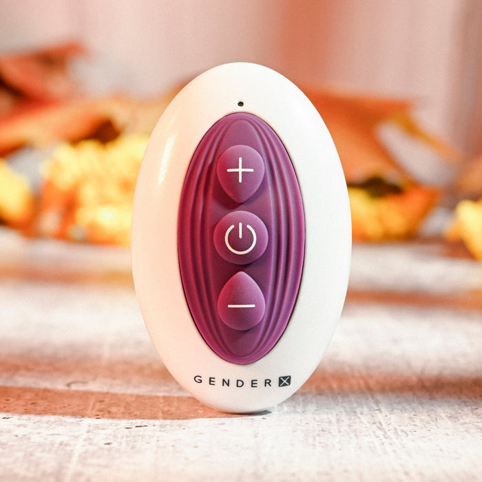 A white oval-shaped silicone vibrator with a purple central section containing three buttons: a plus symbol at the top, a power symbol in the center, and a minus symbol at the bottom. The word "GENDERX" is printed at the base. Against a blurred background of warm colors, this device is revealed to be "The Velvet Hammer Thrusting Hands-Free Dual Stimulation Vibrator" by Evolved Novelties.