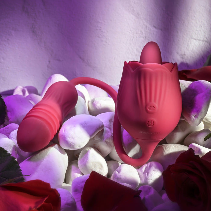 A red, flower-shaped silicone sex toy called "The Wild Rose Licking Tongue and Thrusting Bullet Vibrator" by Evolved Novelties is displayed on white stones, surrounded by red rose petals and illuminated by purple lighting. The toy boasts textured details, a curved, removable attachment, and the tantalizing action of a thrusting bullet vibrator.