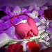 A pink, flower-shaped sex toy, The Wild Rose Licking Tongue and Thrusting Bullet Vibrator by Evolved Novelties, crafted from silicone, is displayed on a white mannequin hand surrounded by white stones and scattered red rose petals. The lighting has a purple hue, highlighting the elegance of this thrusting bullet vibrator.