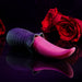 The Tongue Teaser Flickering Realistic Red Tongue Vibrator by Evolved Novelties, featuring a textured black handle, rests on a reflective black surface. In the background, two red roses add a romantic ambiance. The dark lighting highlights the toy and roses, accentuating its promise of realistic oral sensations while being waterproof and submersible.