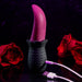 The Tongue Teaser Flickering Realistic Red Tongue Vibrator by Evolved Novelties is shown in a striking scene. The pink, curved adult toy with its textured black base is positioned against a dark background adorned with deep red roses. Connected via a white USB cable, the flexible tongue vibrator exudes hints of realistic oral sensations under a soft purple light.