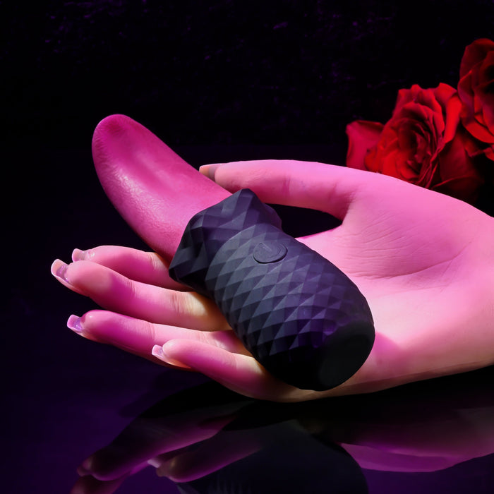 A hand holds the Tongue Teaser Flickering Realistic Red Tongue Vibrator from Evolved Novelties, set against a dark background with a couple of red roses placed to the side. The lighting has a soft pink hue, giving the image a sensual ambiance. This waterproof and submersible device promises realistic oral sensations for an enhanced experience.