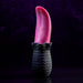 A black, textured base holding a realistic red tongue-like object stands vertically against a dark purple background. The Tongue Teaser Flickering Realistic Red Tongue Vibrator by Evolved Novelties offers lifelike oral sensations, combining the detailed ridged base with the smooth, glossy appearance of the red tongue shape.