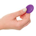 A person's hand holding the Pipedream Products' "Total Ecstasy Remote Controlled Double Penetration Vibrator" between their thumb and forefinger. The small, round purple device, featuring a power button symbol on its surface, resembles a vibrating anal plug. The person's nails are neatly manicured with light-colored nail polish.