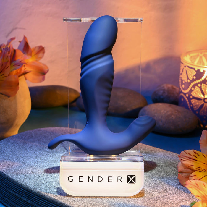 The True Blue Rechargeable Thrusting Silicone Vibrator by Evolved Novelties, an ergonomic adult device designed for ideal g-spot stimulation, is displayed on a clear stand labeled "Gender X." The background features flowers, smooth stones, and a votive candle in a patterned holder, all set against a warmly lit, serene backdrop.