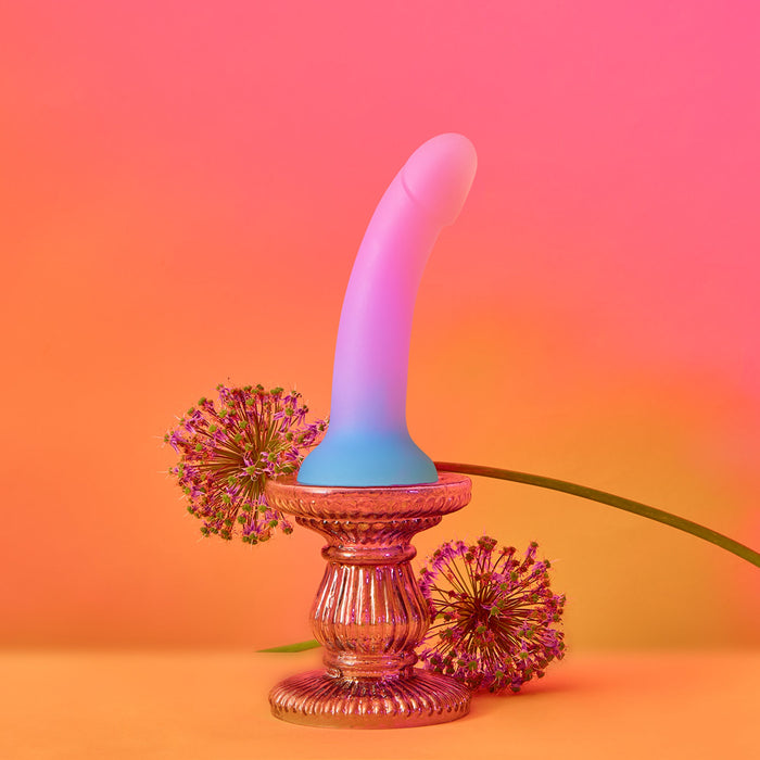 A Utopia Pink and Blue Ombre Silicone Dildo by Lovely Planet is elegantly displayed on a silver ornate pedestal. Surrounding the pedestal are two spherical purple flowers, set against a vibrant gradient background of pink and orange hues.