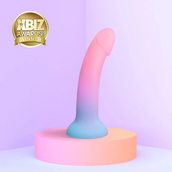 The Utopia Pink and Blue Ombre Silicone Dildo from Lovely Planet stands upright on a circular platform, showcasing its pastel-colored design with a gradient of pink to blue. A gold "XBIZ Awards Winner" badge is displayed in the top left corner against a soft purple background.