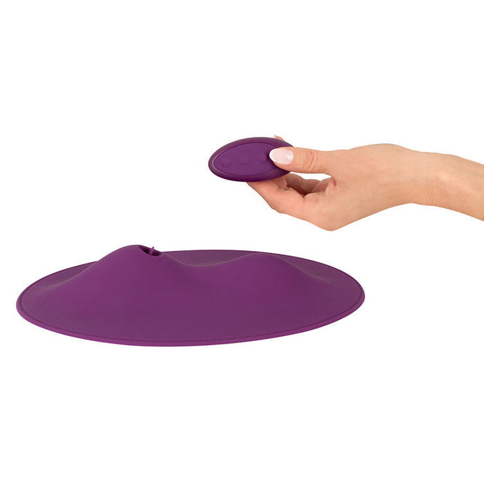 A hand is holding a small, oval-shaped purple remote control above the Orion VibePad 2 Ride On Hands-Free Humping Vibrator with Licking Tongue, a larger, round, and flat waterproof vibrator with a raised center. The VibePad 2 features a sleek design and appears to be made of soft, flexible material.